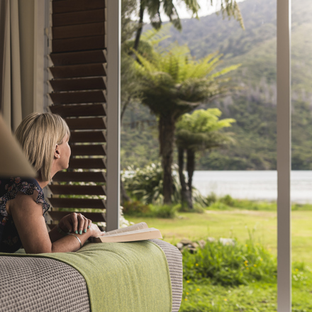 A lady enjoys the water view and a book from an Endeavour Suite at Furneaux Lodge in the Marlborough Sounds, New Zealand.