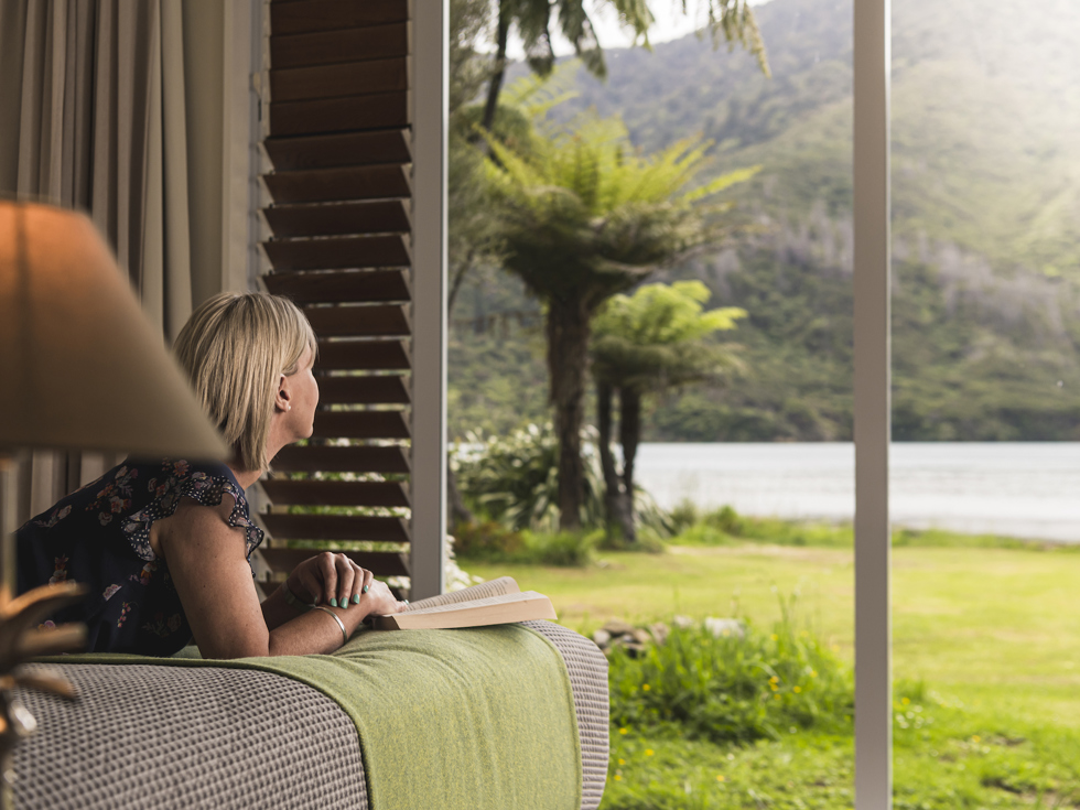 A lady enjoys the water view and a book from an Endeavour Suite at Furneaux Lodge in the Marlborough Sounds, New Zealand.