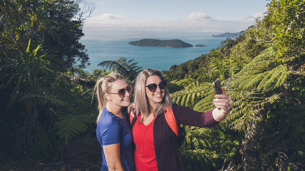 Two women hikers take a selfie photo along Queen Charlotte Track in the Marlborough Sounds, New Zealand, with Motuara Island and sea view in distance.