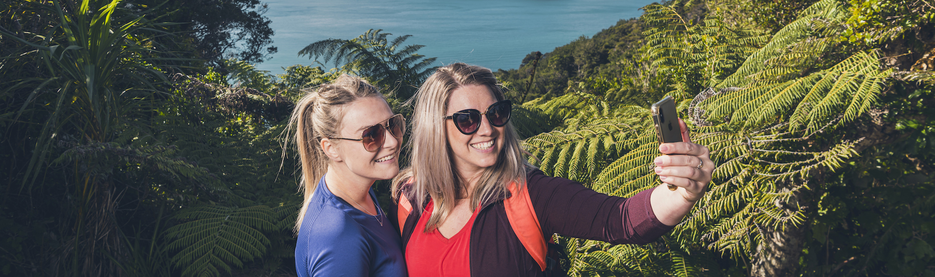 Two women hikers take a selfie photo along Queen Charlotte Track in the Marlborough Sounds, New Zealand, with Motuara Island and sea view in distance.