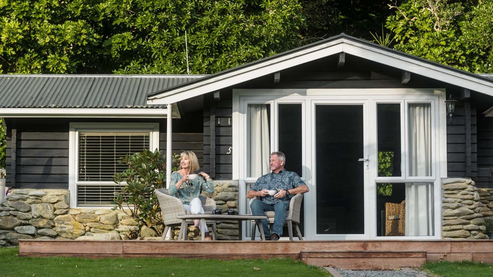 A couple sit on outdoor chairs on their private Cook's Cottage deck with coffee at Furneaux Lodge in the Marlborough Sounds at the top of New Zealand's South Island.