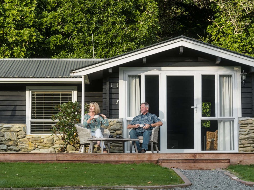 A couple sit on outdoor chairs on their private Cook's Cottage deck with coffee at Furneaux Lodge in the Marlborough Sounds at the top of New Zealand's South Island.