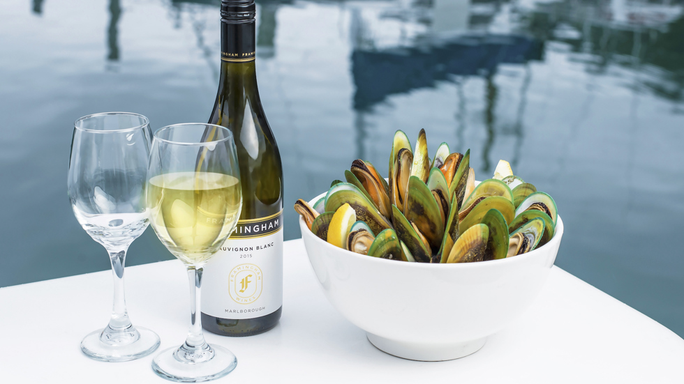 Two glasses of wine, a bottle of wine and bowl of freshly steamed mussels with the Marlborough Sounds in the background, at the top of New Zealand's South Island