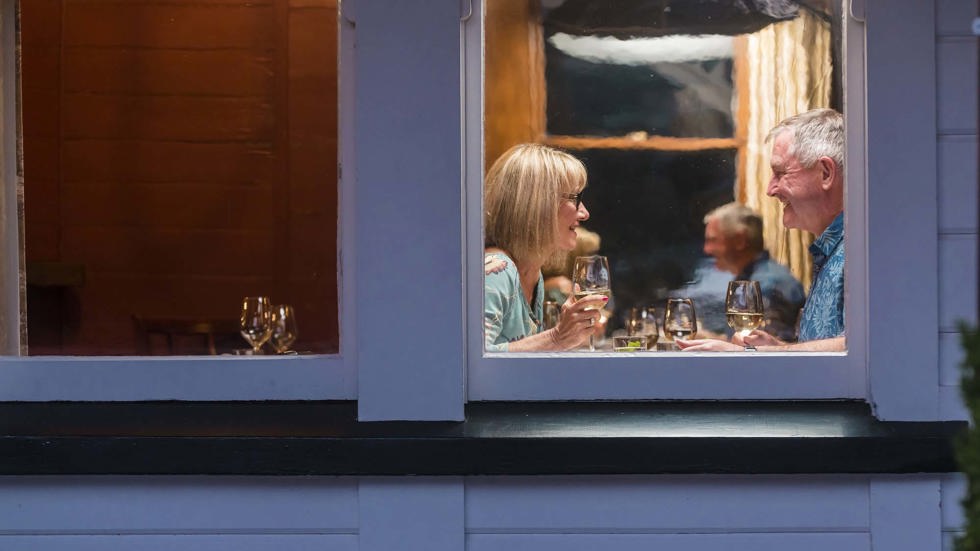 A couple enjoy glasses of local Marlborough wine at a window table during dinner at Furneaux Lodge in the Marlborough Sounds, New Zealand.